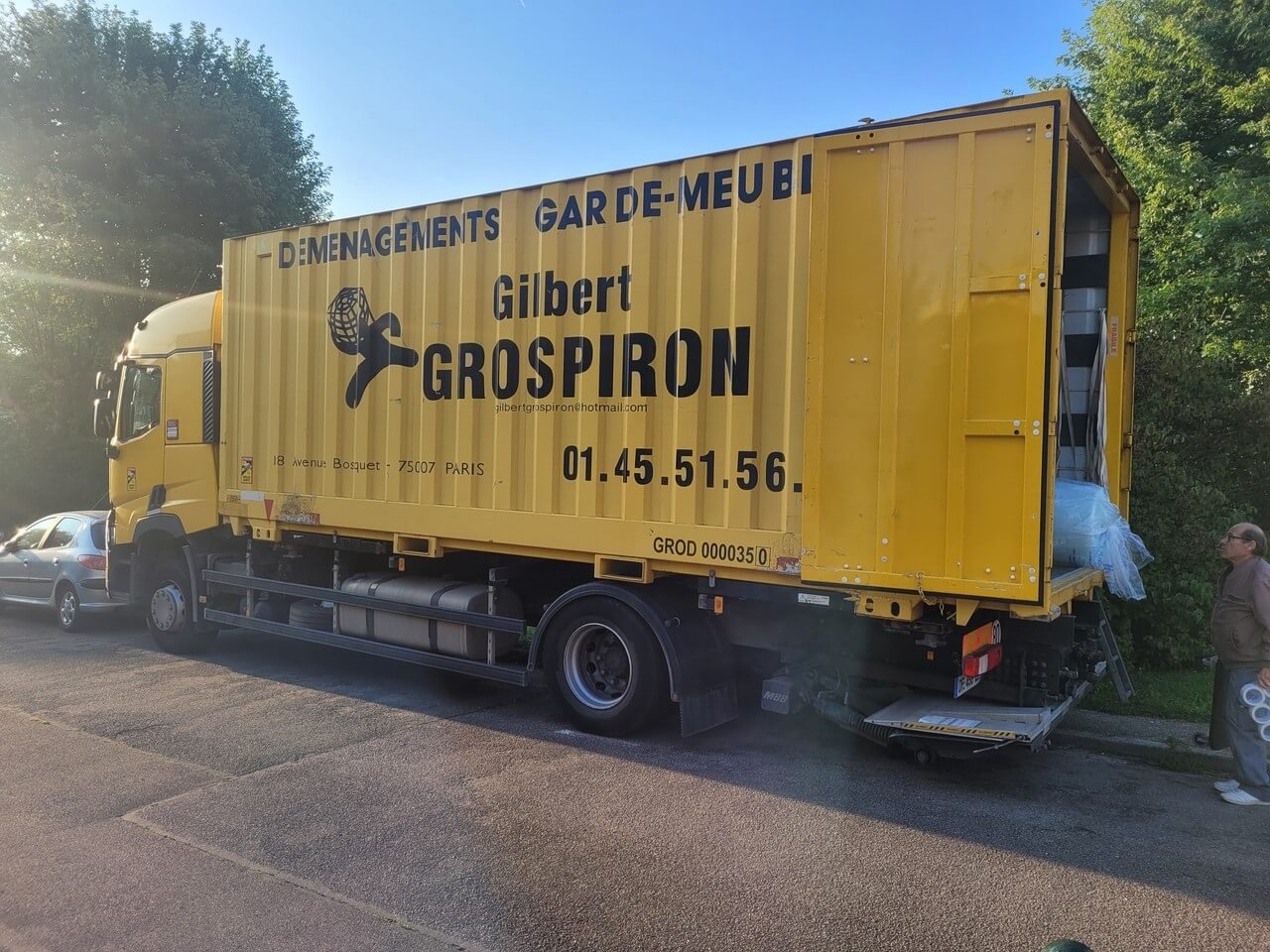 Un grand camion de déménagement jaune de l'agence de déménagement particulier "Gilbert GROSPIRON" est garé sur le bord d'une route, une opération de chargement ou de déchargement en cours.