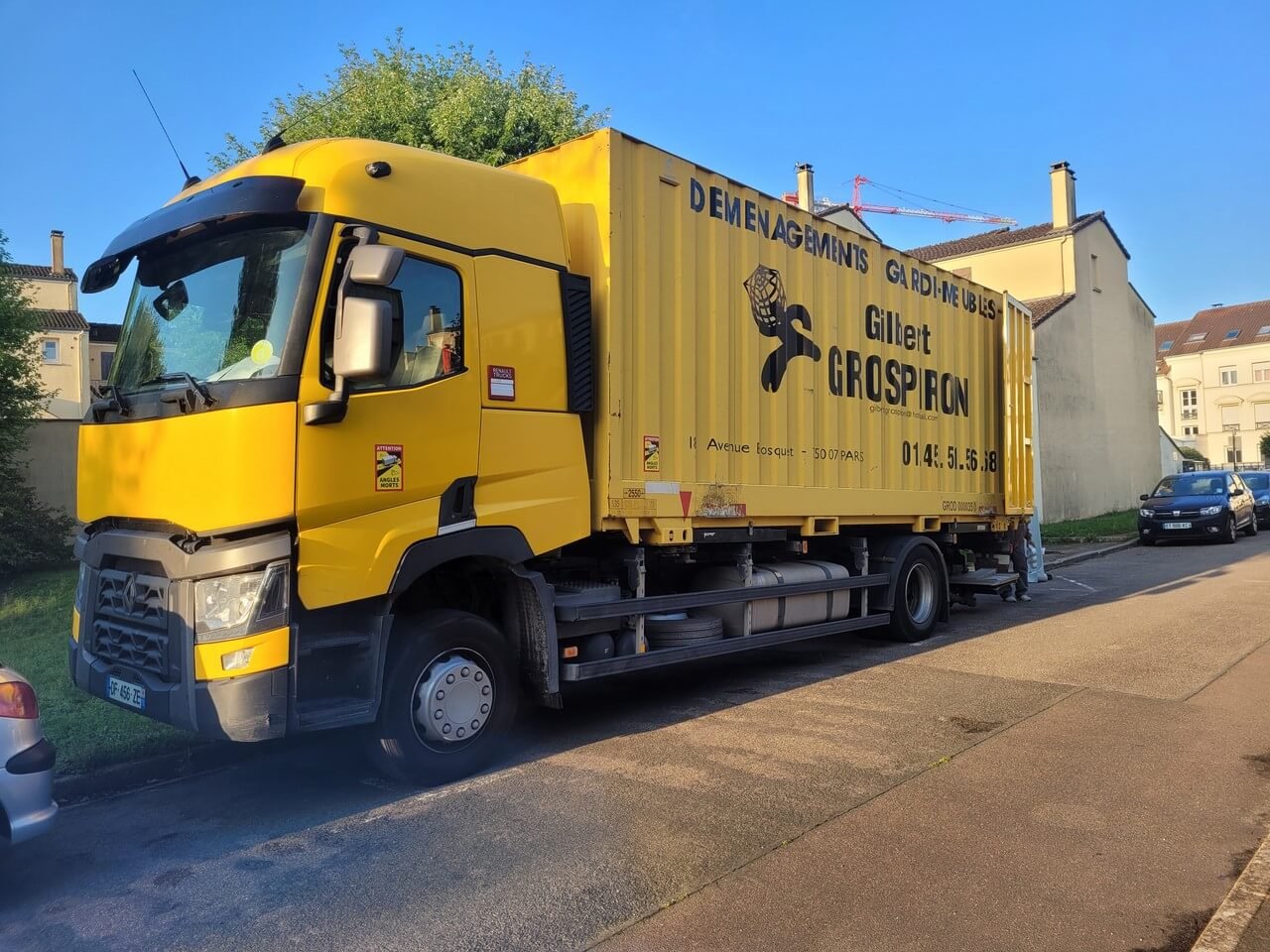 photo camion de déménagement jaune vif, avec l'inscription "DÉMÉNAGEMENTS GARDE MEUBLES Gilbert GROSPIRON", est stationné sur le côté d'une rue. Le soleil illumine le véhicule et les bâtiments résidentiels en arrière-plan, suggérant une journée claire.