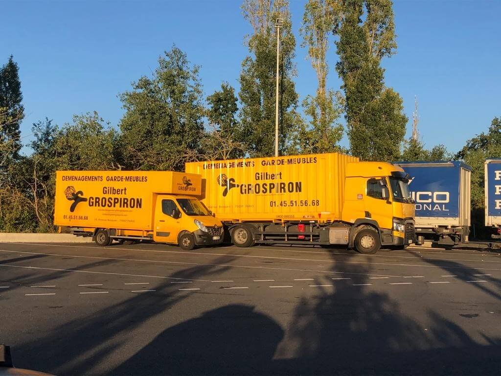 Deux camions de déménagement jaunes de l'agence "Gilbert GROSPIRON" sont garés. À gauche, une plus petite camionnette est stationnée à côté d'un grand camion, les deux affichant le logo et les coordonnées de l'entreprise de déménagement garde meuble